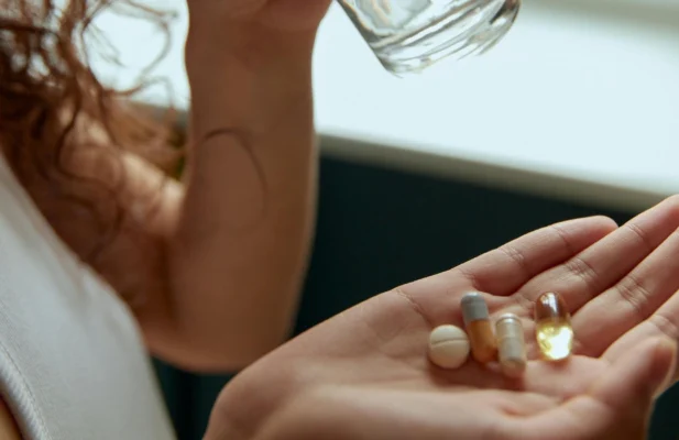 Frau hat auf der Hand mehrere Kapseln und Tabletten und schluckt eine runter mit einem Glas Wasser.